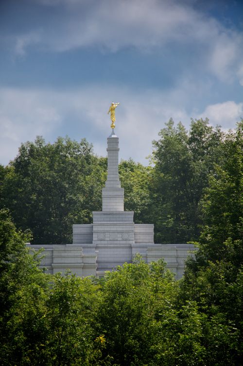 The spire and angel Moroni on the Palmyra New York Temple, seen over the green leaves of the trees on the grounds of the temple, with a blue sky above.
