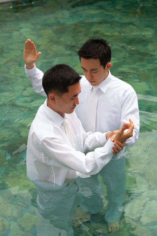 A young man in white clothing being baptized by another young man in an outdoor pool of clean water with rocks seen at the bottom.