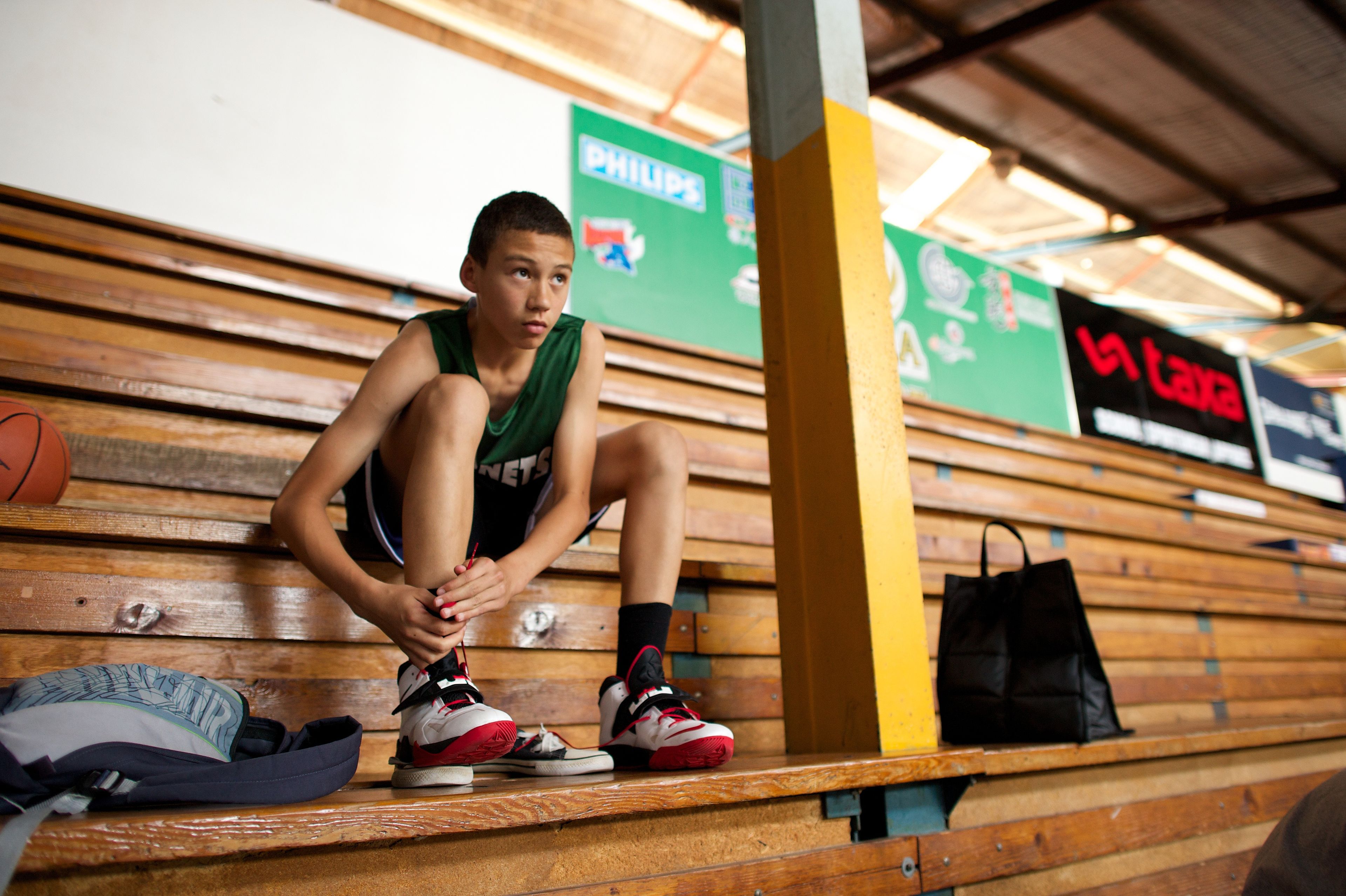 A young man in a basketball uniform sits on the bleachers, warming up as he prepares to go out on the basketball court.