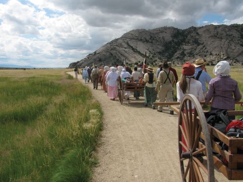 A large group of men and women, all dressed as pioneers, walk down a road past a mountain, pushing and pulling handcarts full of equipment.