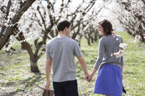 A young couple holding hands and walking through an orchard with blossoms.