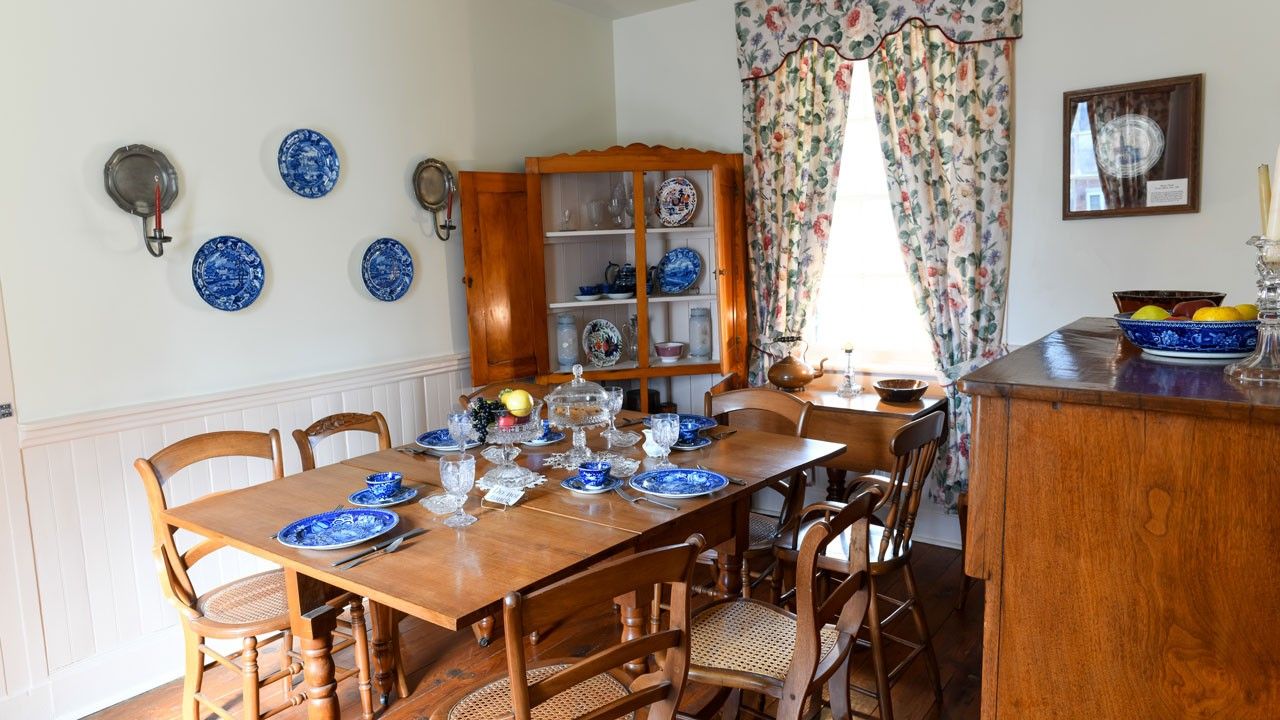 A table set with blue china surrounded by chairs. Three blue china plates are hanging on the wall, and a hutch in the corner of the room is open to display plates and vases.