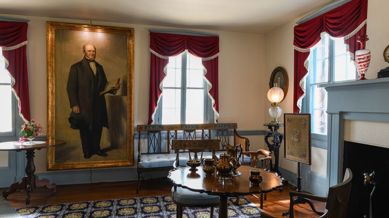 Interior view with life-size portrait of a man on a wall, a tea set on a table with chairs in the foreground, and red-curtained windows in the background.