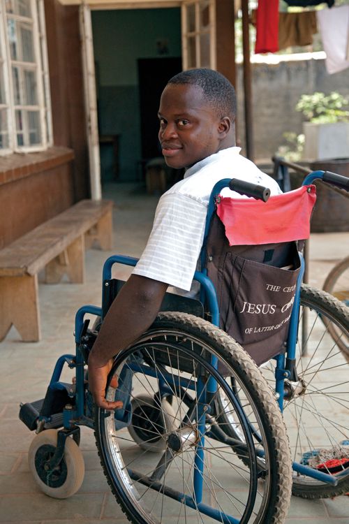 A man from Africa in a light blue striped shirt, smiling and sitting outside in a wheelchair, resting his hand on a wheel.