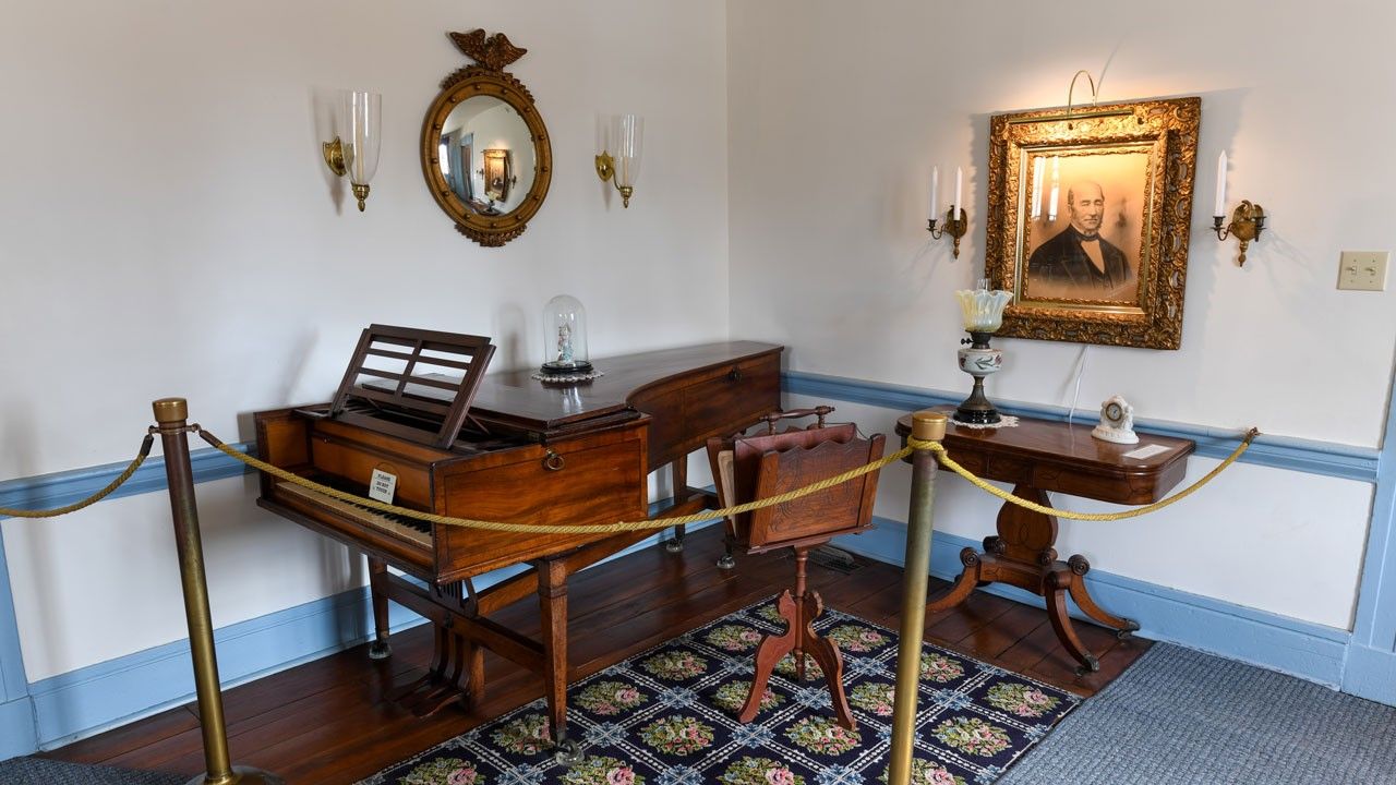 A baby grand piano behind roped stanchions in a room with white walls, light blue trim, and a floral rug.