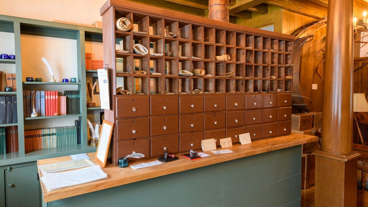 Close-up showing large wooden post office box resting on a cabinet painted green. Shelves holding books and paper are in the background. 