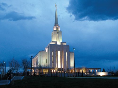 The Oquirrh Mountain Utah Temple, with the lights on in the evening, showing a short set of steps leading toward the temple on the left side.