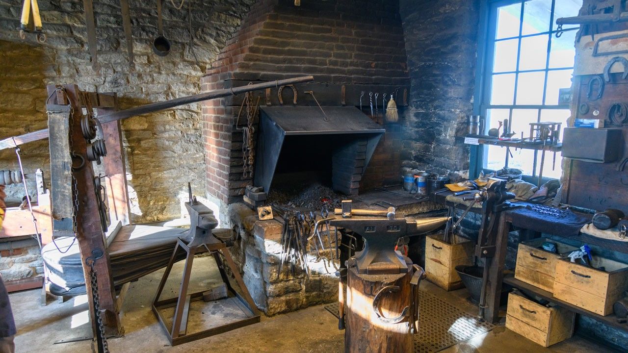 Interior view of a blacksmith shop with an anvil on a stump in the foreground, a forge, bellows, and many tools and horseshoes all around.