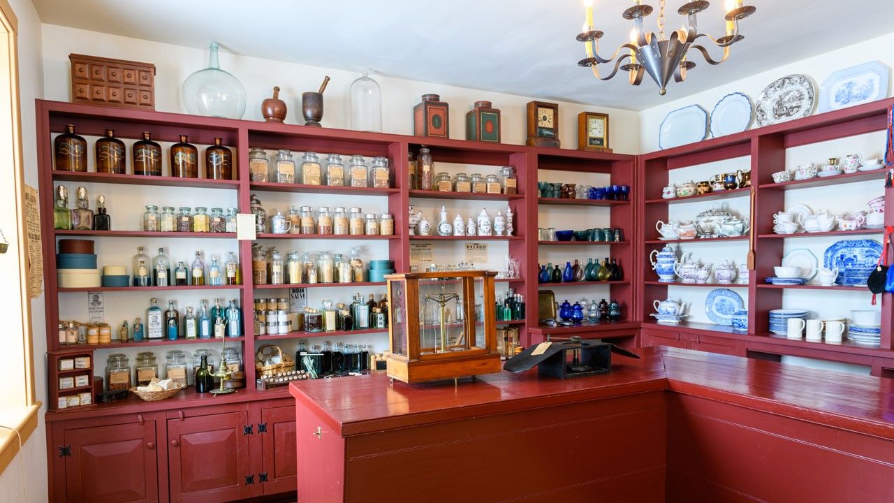 A drug store with a red counter and red shelves filled with ninteenth century apothecary bottles and jars, as well as patterned china.