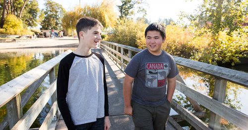 Zwei Jugendliche gehen über eine Brücke. Es ist Herbst. Die Jungen unterhalten sich. Die Brücke führt über einen See. In der Ferne sieht man Felder und Spielgeräte.