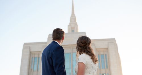 Young couple stands outside the Ogden Utah Temple. They are looking out at the temple in the background with the Angel Moroni being almost top center of the image. He is wearing a dark blue suit with white collared shirt. She is in a white dress. It is late afternoon/early evening.