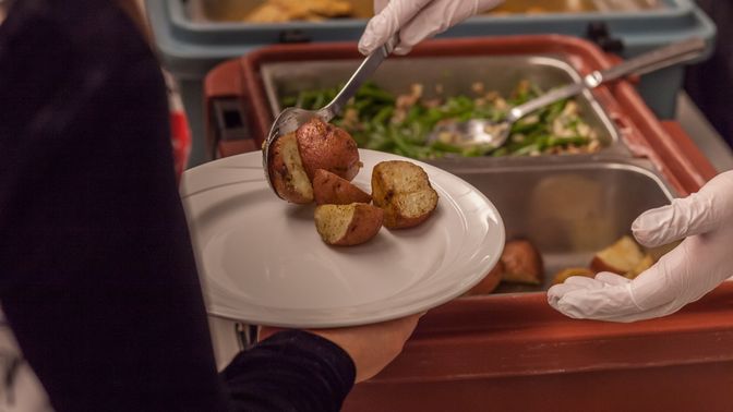 A person holding out a plate is served roasted potatoes at a buffet style meal.