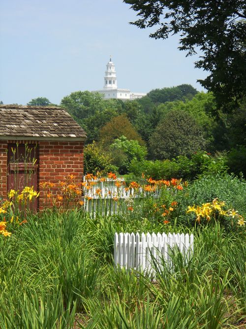 The Nauvoo Illinois Temple seen from afar at the top of a hill, with green vegetation and a small red brick building in the foreground.