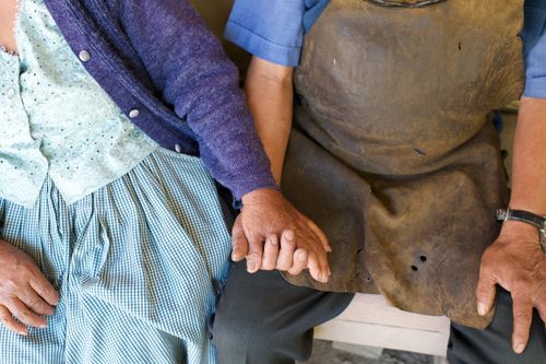 A woman in a blouse, sweater, and plaid skirt holding hands and sitting beside her husband in a blue shirt, work apron, and watch.