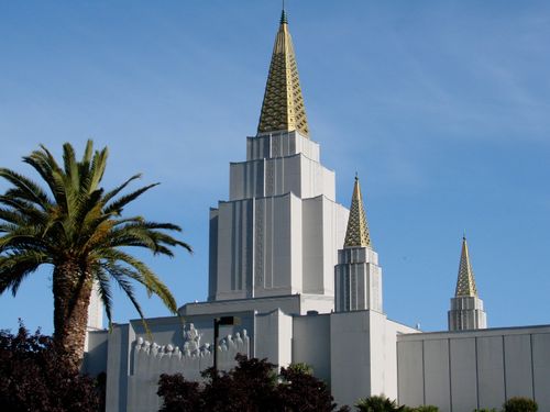 Three of the spires on the Oakland California Temple on a bright day, with a clear blue sky above and a large palm tree in the front.