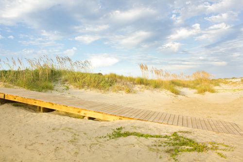 A wooden boardwalk leading through the sand and brush, with clouds in the sky.