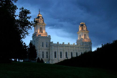 The Manti Utah Temple lit up in the late evening, behind silhouetted trees and bushes.