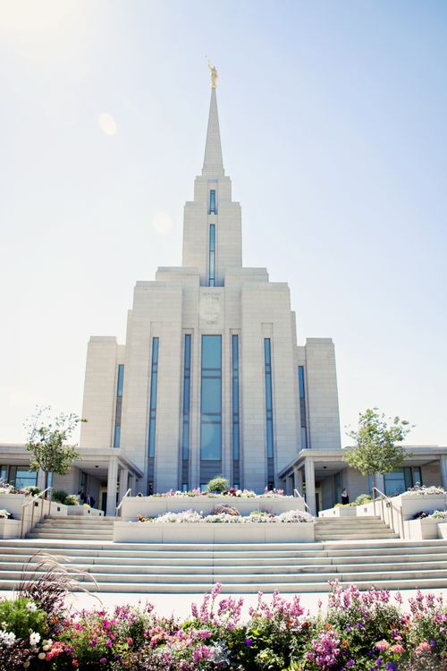 A front view of the Oquirrh Mountain Utah Temple on a spring day, with flowers and plants in the flower beds near the temple steps and door.