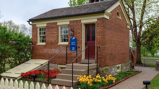 Small red brick building with concrete steps leading into the home. There are flowers blooming in the dooryard garden.