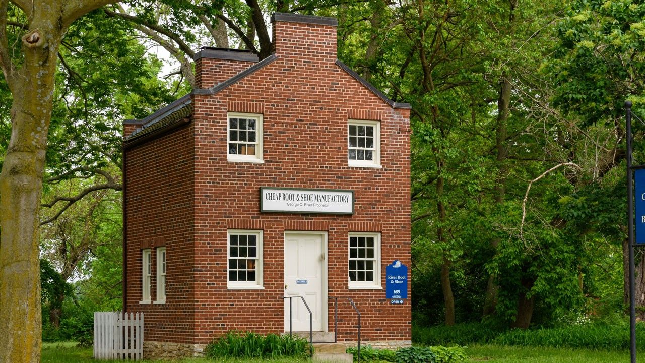 Two-story red brick building with a sign reading “Cheap Boot & Shoe Manufactory."