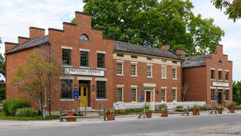 A complex of three two-story red brick buildings. The building on the left has a sign that reads “Printing Office." The building on the right reads “Post Office."