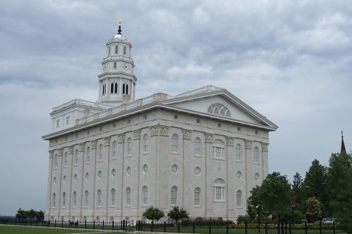 The back and side of the Nauvoo Illinois Temple, with a pale blue and clouded sky overhead and the temple grounds in view.
