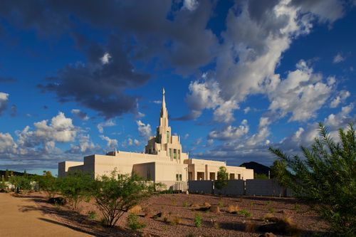 A side view of the Phoenix Arizona Temple at sunrise, with small gray and white clouds overhead and desert plants in the foreground.