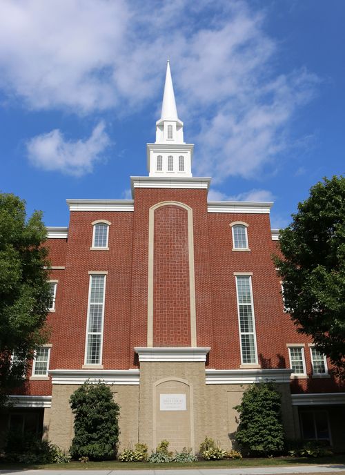 A front view of a tall, red-brick chapel with a white steeple and trees on each side in Cambridge, Massachusetts.
