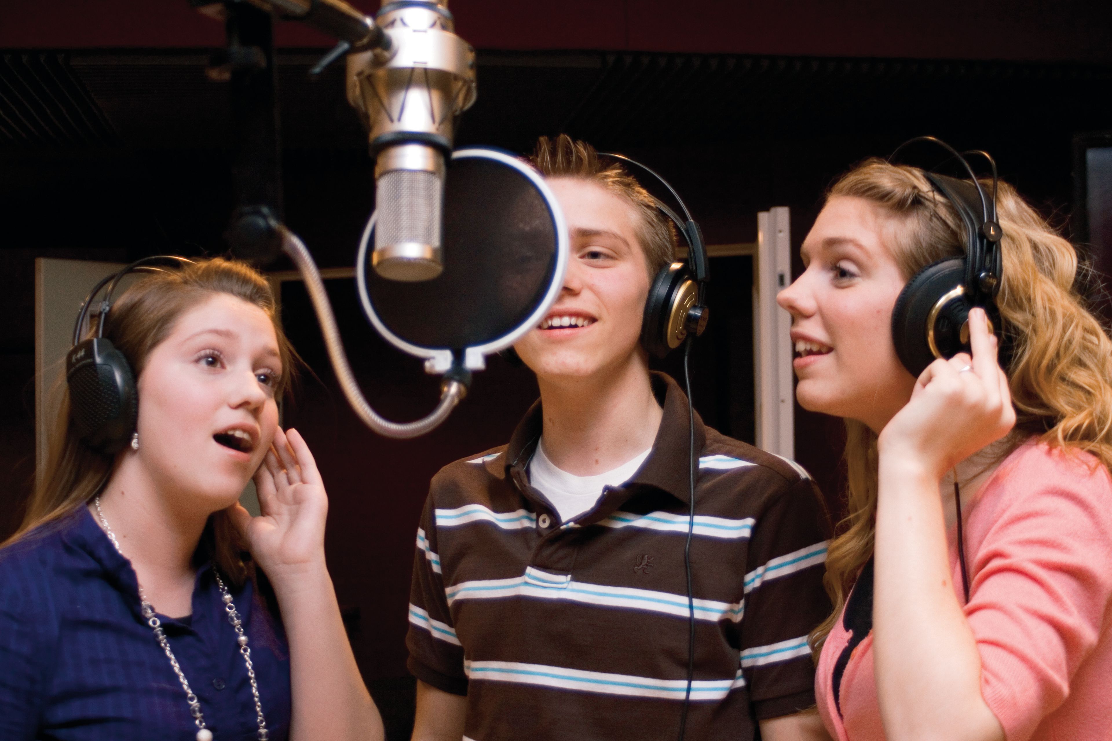 Three youth stand around a microphone together and sing in a recording studio.