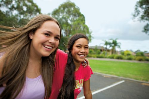 A young woman with long brown hair and a purple shirt puts her arm around another young woman with long dark brown hair and a T-shirt as they walk together and smile.