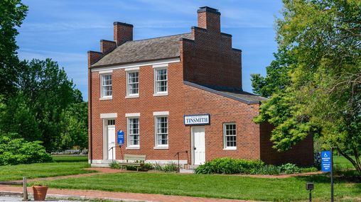 Two-story red brick building with a lean-to wing on the north side with a sign that reads “Tinsmith."