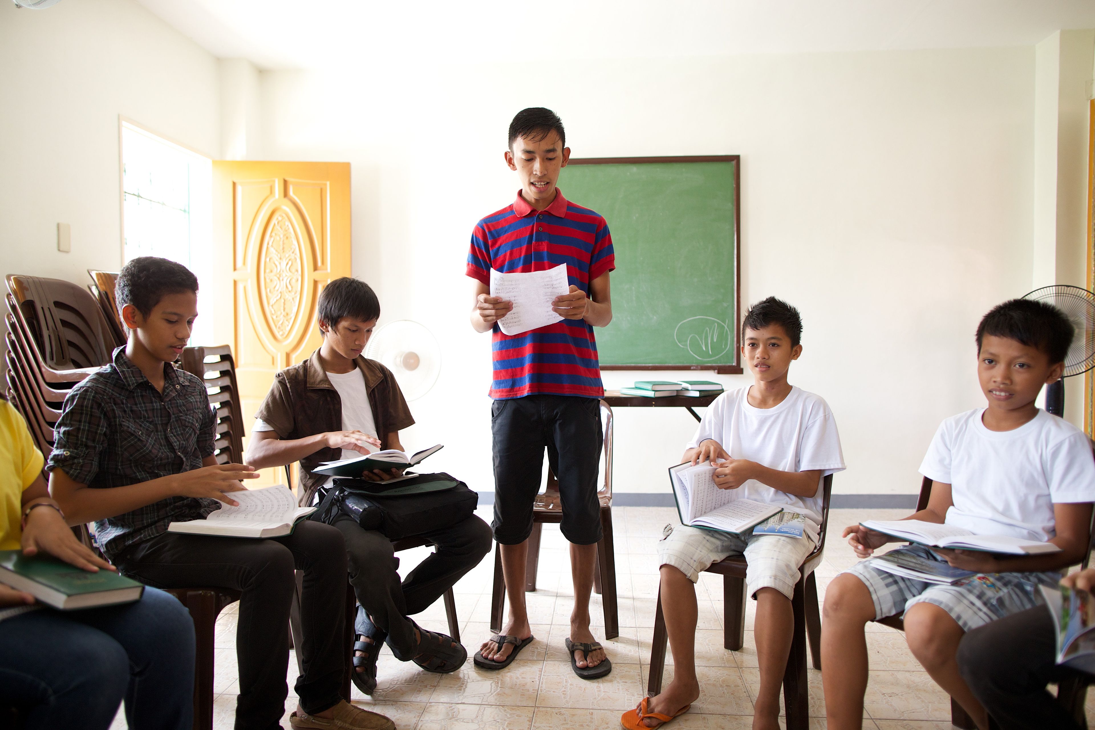 A group of young men from the Philippines sitting in a circle while one young man stands and reads to them.