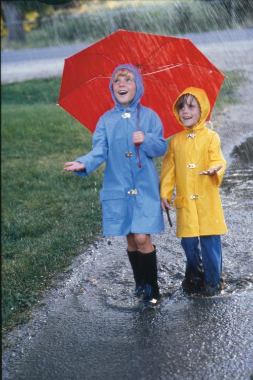 A girl in a blue raincoat holds a red umbrella and plays in the rain with a girl in a yellow raincoat.