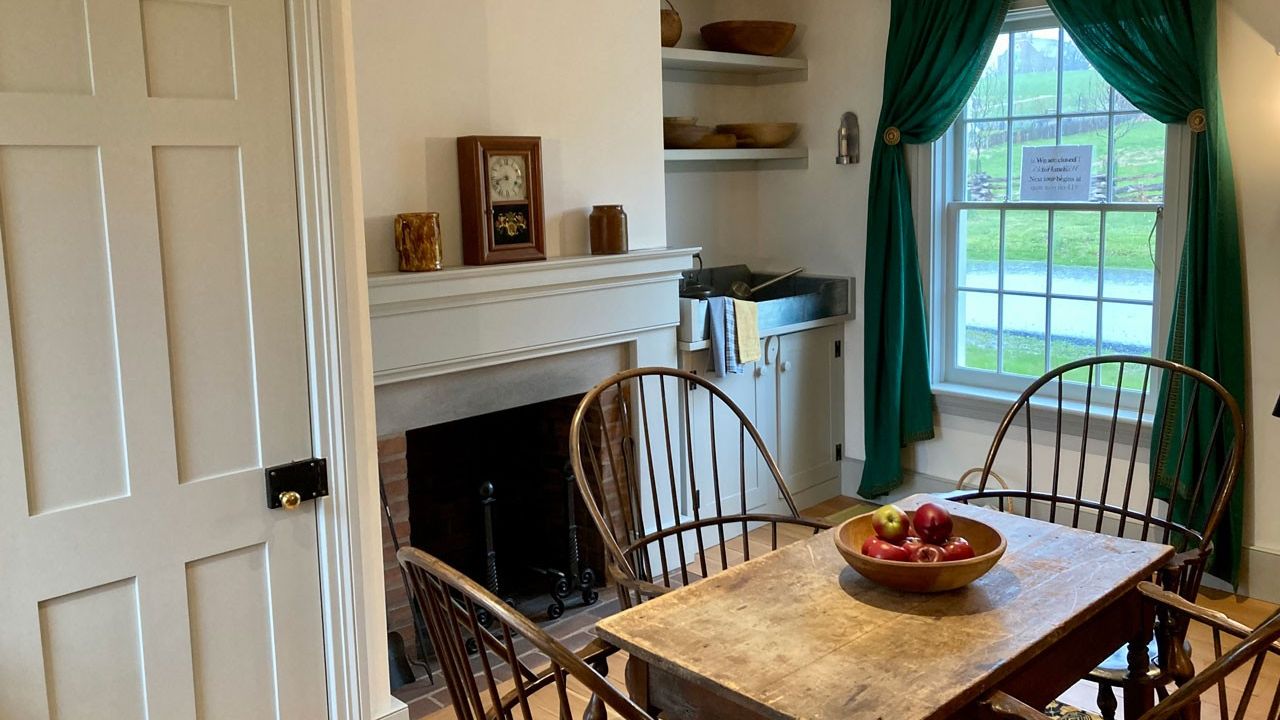 A dining room with four chairs surrounding a table with a bowl of fruit on it in. A window with green curtains is in the background.