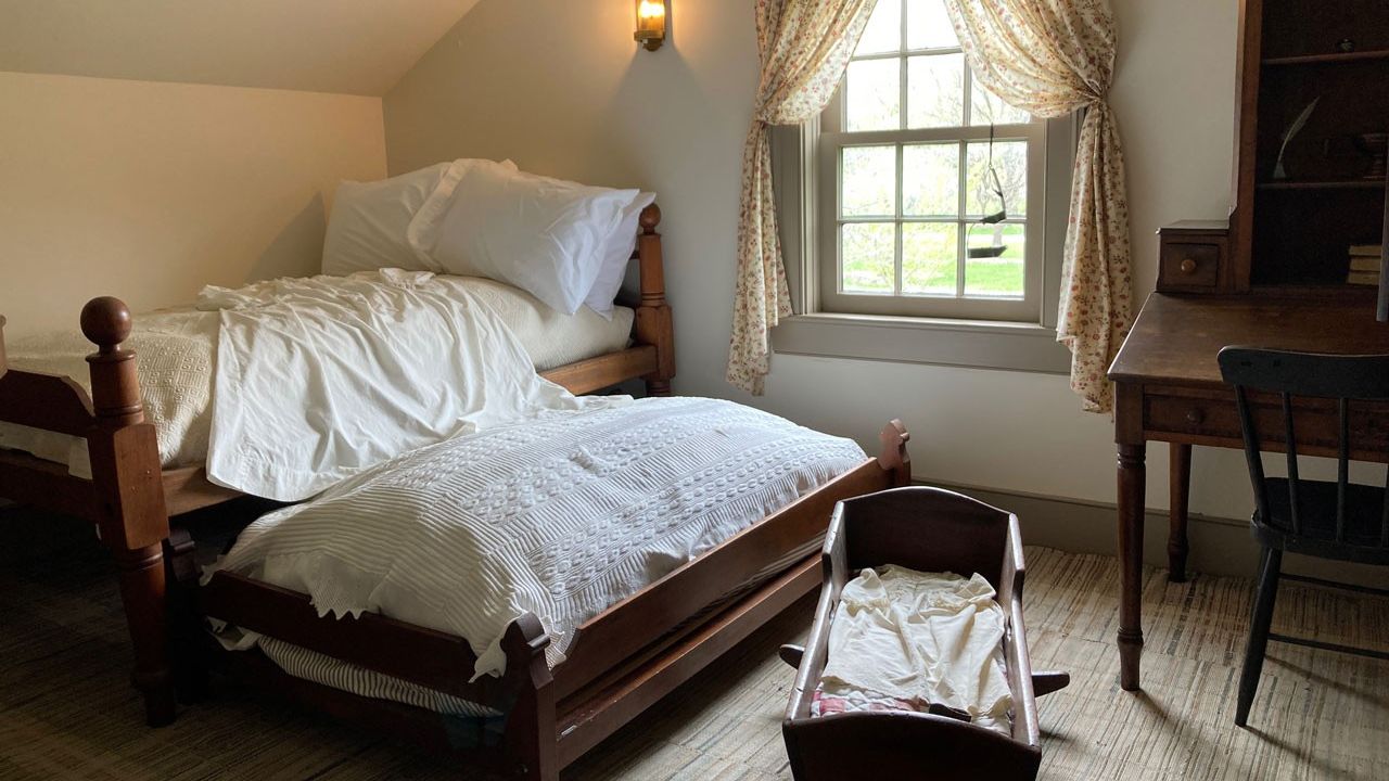 A bedroom with a bed, pullout bed, and cradle. A desk is next to the window against the wall.