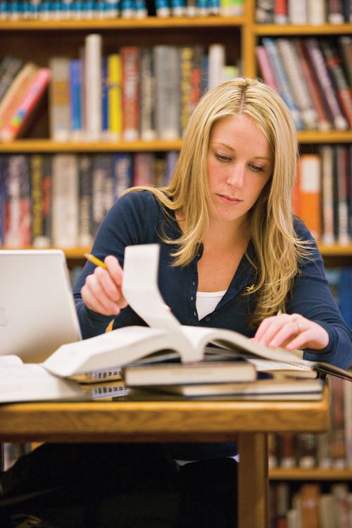 A young woman with long blonde hair and a blue shirt sits at a table in a library and flips through a book, with a bookshelf in the background.
