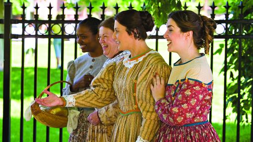 Four women standing on a street next to each other dressed in pioneer clothing.