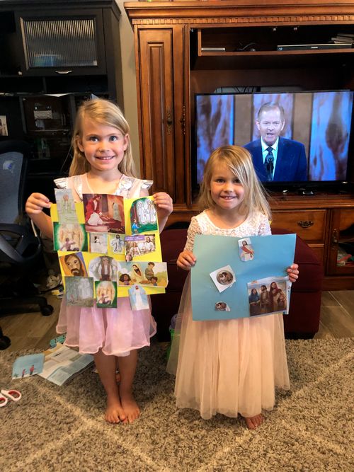 Lily and Avery Collette stand in front a television and hold up conference collages while Elder Gary E. Stevenson speaks on the TV behind them.