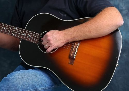 A close-up image of a man in a black shirt and jeans sitting down and strumming the strings on a guitar.