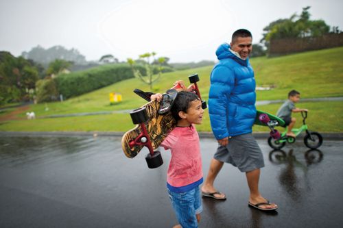 A father plays with two sons outside in the rain. One is skateboarding; the other son is riding a bike.