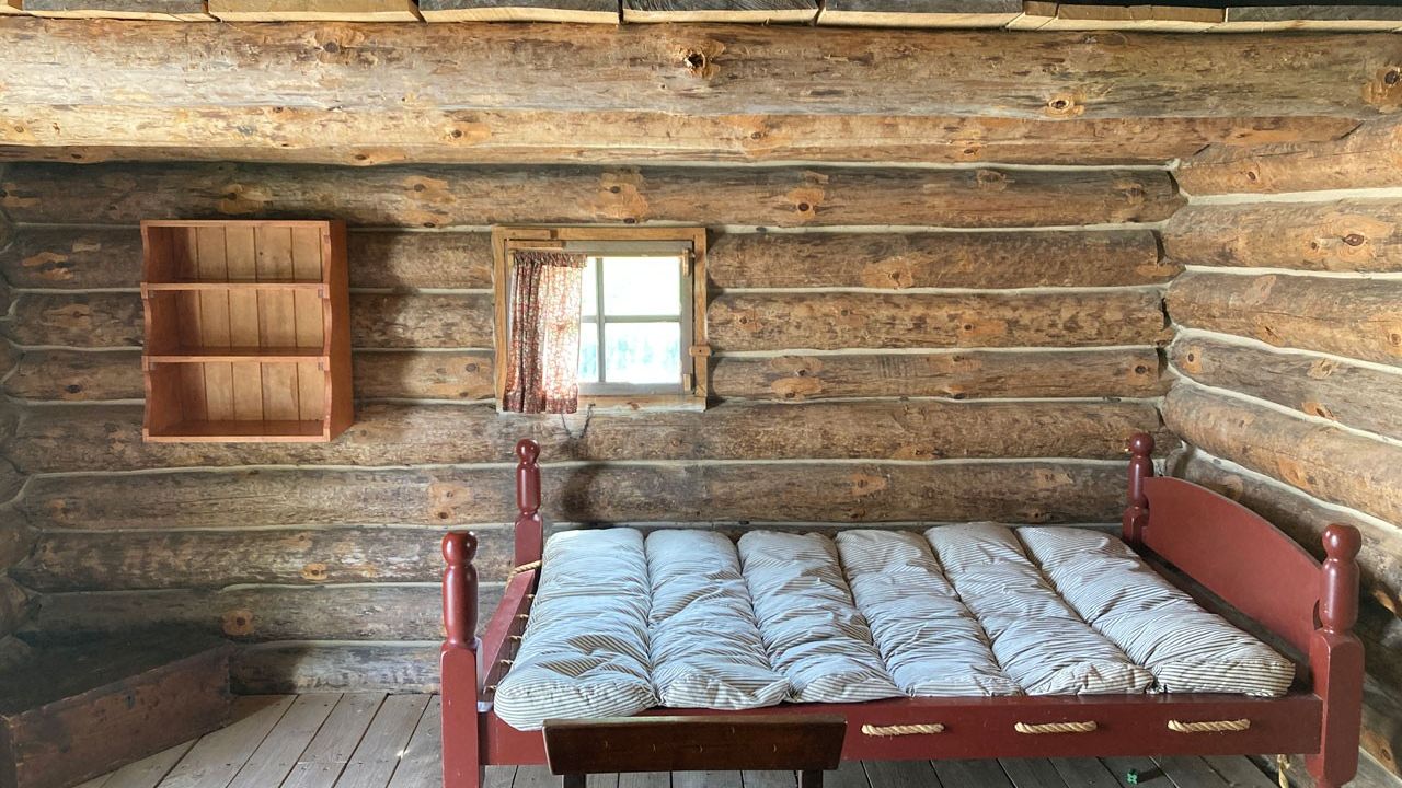 Interior of a log cabin sparsely furnished with a bed, empty shelving, and wooden trunk.