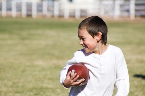 A boy with dark brown hair and a white shirt smiles while holding a football on a grass field.