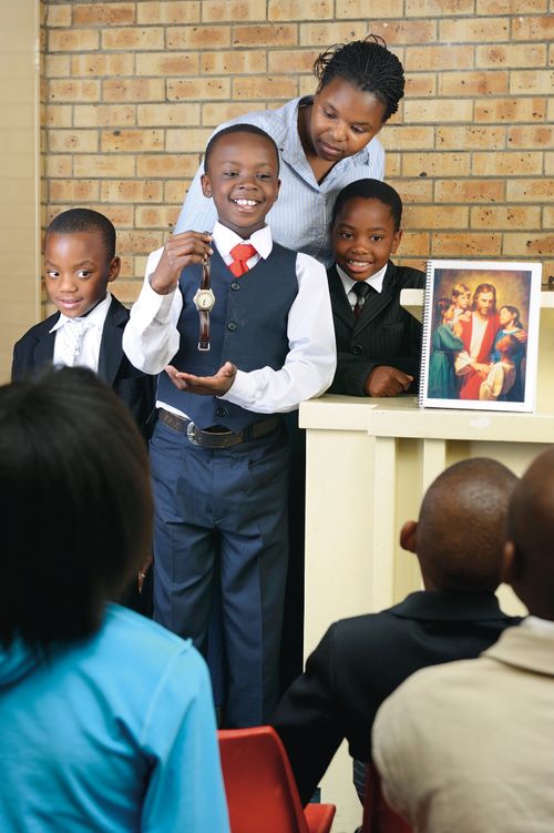 A young boy in a vest, tie, and slacks shows a watch during sharing time while standing beside two boys and his teacher.