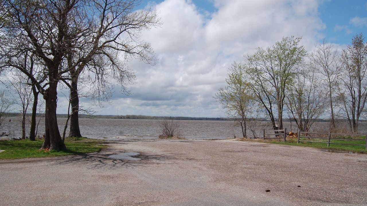 An asphalt road ending at the Mississippi River with trees on either side of the road and large clouds in the blue sky above.