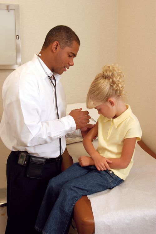 A doctor in a white shirt and black pants giving an injection in the arm of a young girl with blonde curly hair sitting on a bed.