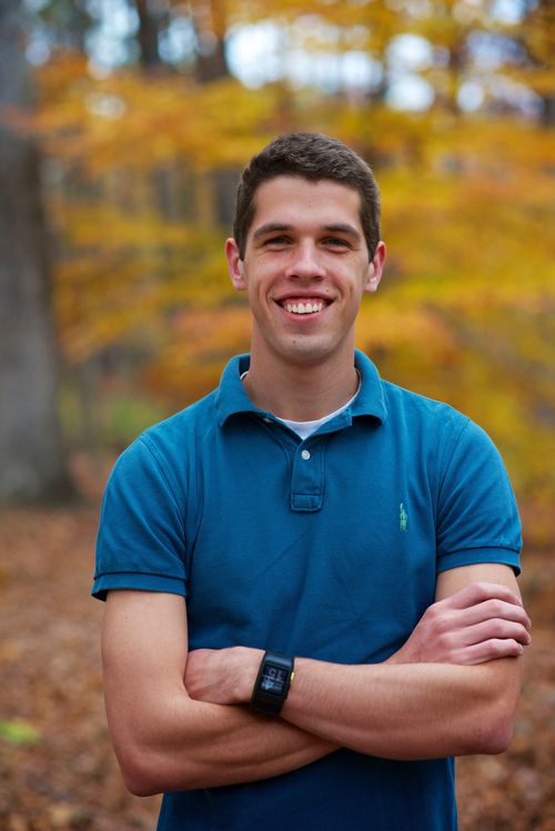 A portrait of a young man in a blue polo shirt with his arms folded, standing outside in the fall and smiling.