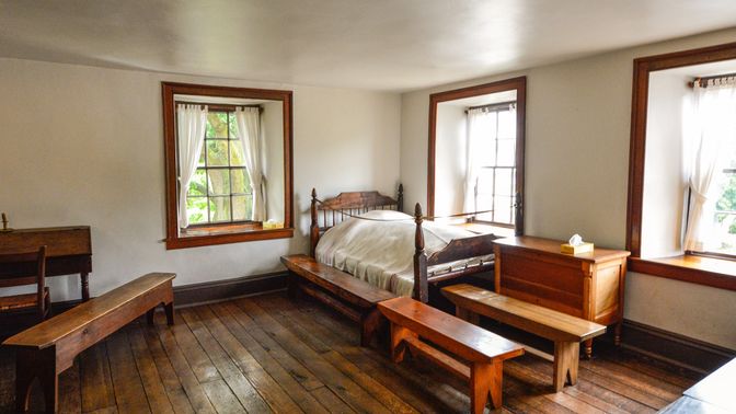 View of a room with a bed in the corner, benches at the foot of it, and a desk to the left.