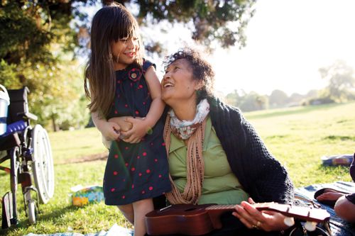 A woman sits outside, holding a ukulele in one arm and hugging her granddaughter in the other.