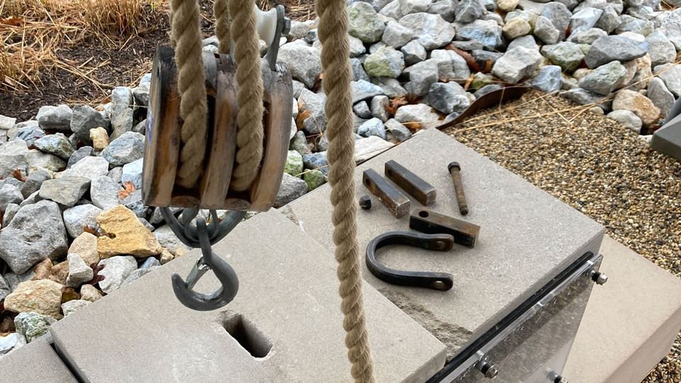 A rope and pulley hang above three stone blocks with a disassembled metal tool strewn on top. 