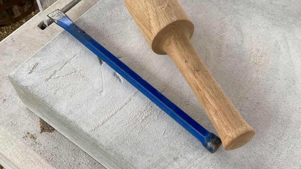 A wooden mallet and metal chisel rest on top of a stone slab and base. 